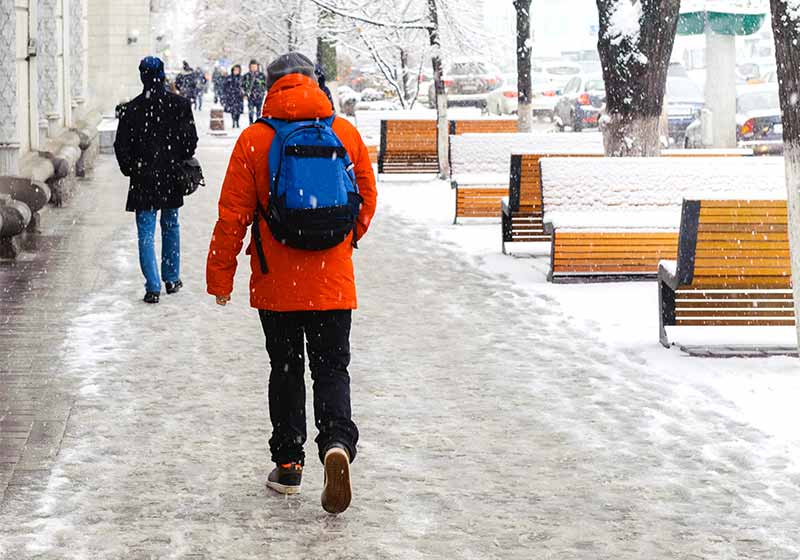 A young man in a red jacket with a blue backpack walks on a snow-covered sidewalk during a snowstorm | winter walking shoes