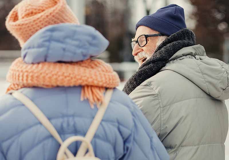Elderly man in winter coat and hat smiling to his wife while walking on winter day with her | winter walking tips