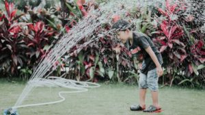 Boy standing under garden sprinkler | How To Save on Water Bill In Smart Ways | Featured