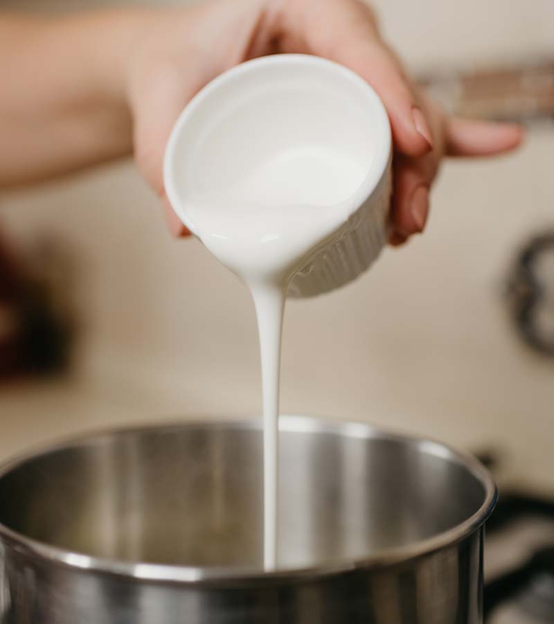 A close photo of a hand of a woman who is pouring cream from the cup to the saucepan | how to make caramel sauce