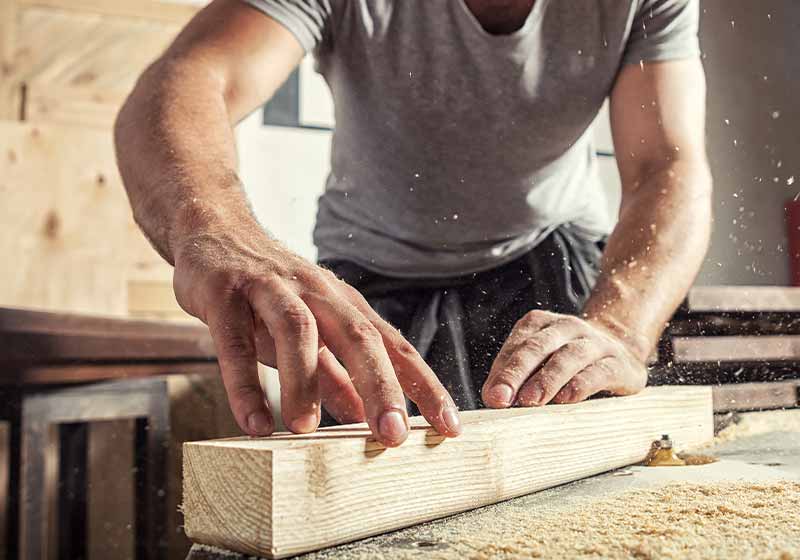 A young male carpenter builder in a gray T-shirt and working overall equals a wooden bar with a milling machine in the workshop | rabbit farm